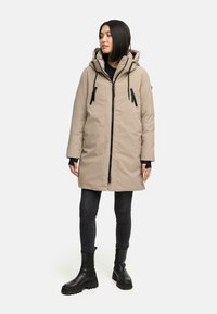 Beige lang parka med hætte, sort lynlås og snore. Båret over en sort sweater og parret med mørke jeans og sorte støvler.