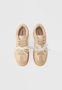 Μπεζ σουέτ αθλητικά παπούτσια με καουτσούκ σόλες, μπεζ κορδόνια και διακοσμητικά λευκά δαντελένια και μαργαριταρένια φιόγκια δεμένα από πάνω, απεικονιζόμενα από πάνω σε λευκό φόντο.