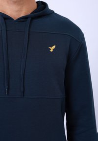 Tamno plava hoodie s teksturiranim završetkom, s kanapima i malim zlatnim logotipom ptice na prsima. Ima dizajn s vertikalnim šavovima.