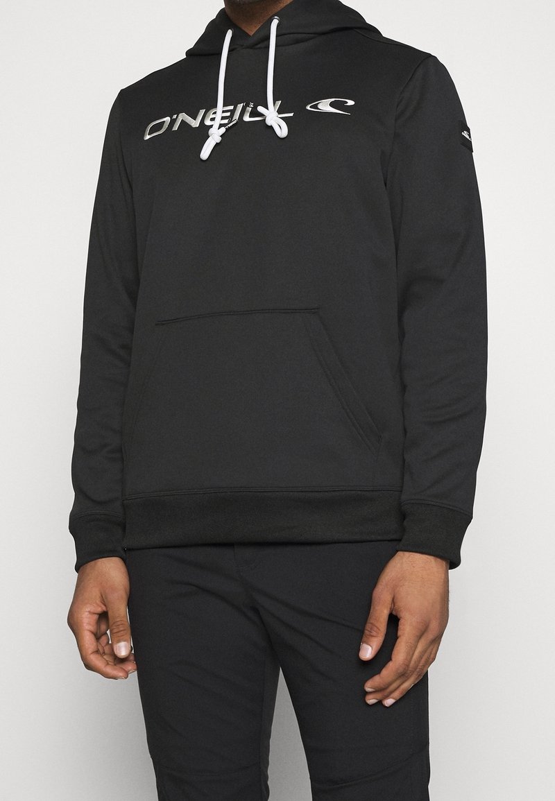 Personne portant un sweat à capuche noir O'Neill avec des cordons blancs et un pantalon noir, debout devant un fond clair uni.