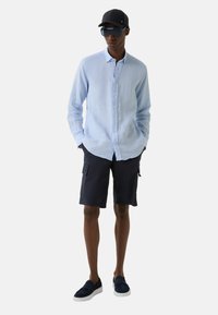 Lichtblauwe lino knoopshirt, marineblauwe cargoshorts, zwarte pet, grote blauwgetinte zonnebril en marineblauwe instappers.