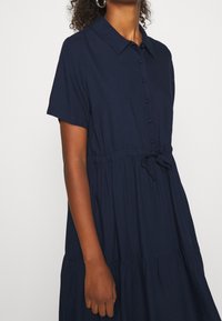 Navy blue sukienka z krótkim rękawem, z kołnierzykiem, zapinana na guziki z przodu, z elastycznym pasem i warstwowym dołem, wykonana z miękkiego, lekkiego materiału.