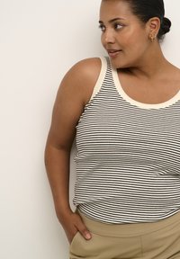 Gestreifter Tanktop in Schwarz und Weiß mit einem cremfarbenen Ausschnitt; gerippte Textur; getragen mit beigen Hosen; das Modell posiert seitlich vor einem hellen Hintergrund.