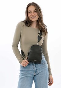 Černá kožená crossbody taška s zaobleným víkem, stříbrným kováním a nastavitelným popruhem, nošena přes béžový dlouhý rukáv.