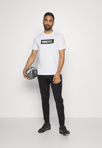 Weißes Nike F.C. T-Shirt mit schwarzem Logo, schwarze Sweatpants und schwarze Schuhe, hält einen schwarzen Fußball mit demselben Logo.