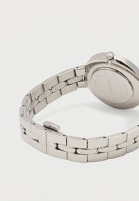 Ασημένιο μεταλλικό μπρασελέ ρολογιού με ανοιχτό κούμπωμα, που δείχνει την πίσω πλευρά της στρογγυλής θήκης ρολογιού χαραγμένη με λεπτομέρειες μάρκας και υλικού.