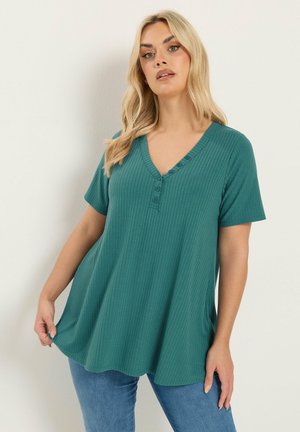 Frau trägt ein türkisfarbenes, geripptes Kurzarmoberteil mit Knöpfen, kombiniert mit blauen Jeans, steht vor einem schlichten hellen Hintergrund.