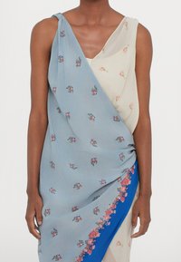 Robe bleu et crème présentant des motifs floraux, un design drapé, un côté noué, et des accents bleus le long de l'ourlet. Fabriquée en tissu léger.