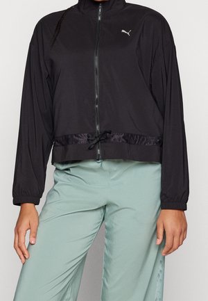 Henkilö, jolla on yllään musta Puma-vetoketjullinen takki, jossa on kiristysnauha vyötäröllä, ja vaaleanvihreät housut, seisoo yksinkertaisen taustan edessä.