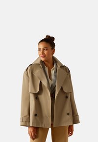 Beige double-breasted trench coat with large lapels, button accents, and a relaxed fit. Worn over a checkered shirt and belted trousers.