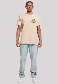 Beige cotton t-shirt with a black graphic design featuring a grill and the words "Grill Master Dad." Worn with light blue jeans and white sneakers.