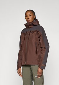 Giacca impermeabile marrone e grigia con cappuccio, zip frontale, tasche con zip e dettagli con logo. Tessuto testurizzato per uso all'aperto.