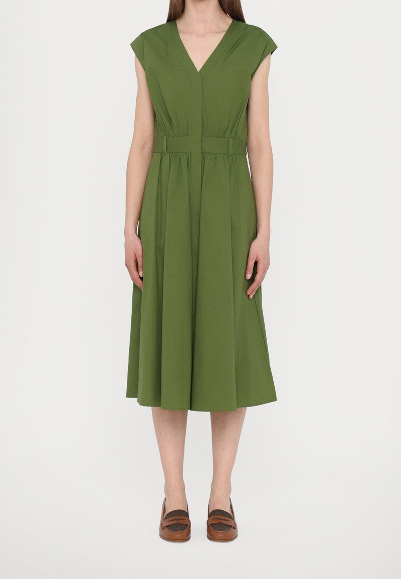 Femme portant une robe midi sans manches vert olive avec un col en V et une taille cintrée, associée à des chaussures plates marron.