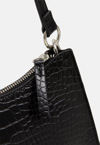 Mustan krokotiilitaskukkaan kulma, jossa on hopeinen vetoketju, metallirenkaat ja kiinnitetty hihna valkoista taustaa vasten.