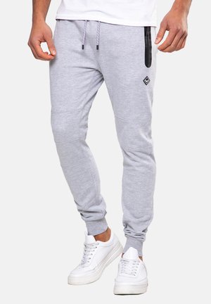 Homme portant un pantalon de jogging gris clair avec une poche zippée noire, des baskets blanches et une chemise blanche, debout devant un fond blanc.