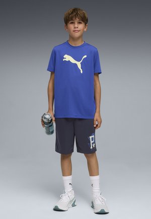 Niño de pie, vistiendo una camiseta azul Puma, pantalones cortos oscuros, calcetines blancos Puma y zapatillas deportivas, sosteniendo una botella de agua en su mano derecha.