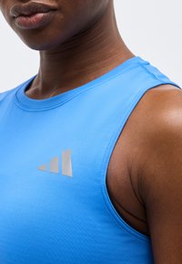 Άτομο που φοράει αμάνικη αθλητική μπλούζα σε ανοιχτό γαλάζιο χρώμα με ένα μικρό αντανακλαστικό τριγωνικό λογότυπο στο στήθος, πάνω σε απλό φόντο.