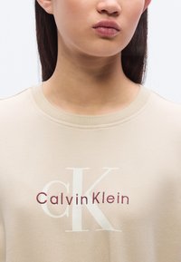 Μπεζ φούτερ με στρογγυλή λαιμόκοψη που φέρει το λογότυπο 'Calvin Klein' σε λευκό και μαρόν. Μαλακή υφή και απλός σχεδιασμός χωρίς πρόσθετα διακοσμητικά.