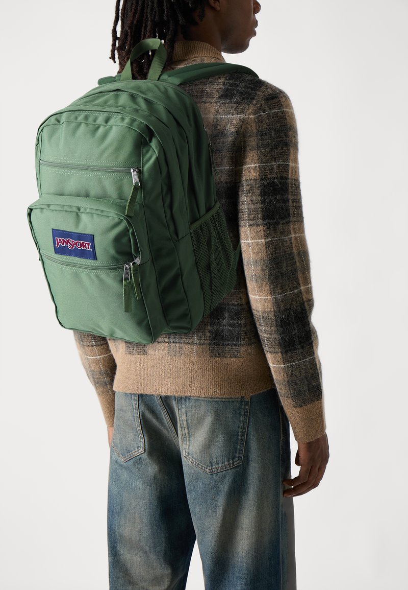 Zöld Jansport hátizsák tartós anyagból készült, több cipzáras rekesszel rendelkezik, és hálós oldalzsebe van. Kockás pulóver felett viselve.