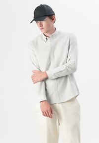 Camisa de botones con finas rayas verticales, mangas largas y un bolsillo. Se lleva con pantalones de color claro y una gorra negra. Textura de tela suave.