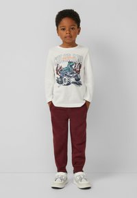 Weißes Langarmshirt mit einem bunten Grafikdruck, der einen Dinosaurier und einen Monstertruck zeigt, kombiniert mit burgunderfarbenen Jogginghosen und hellen Sneakern.