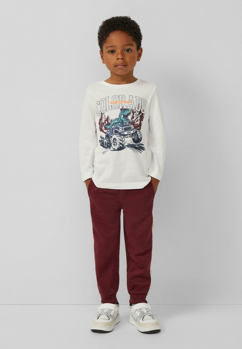 Weißes Langarmshirt mit einem bunten Grafikdruck, der einen Dinosaurier und einen Monstertruck zeigt, kombiniert mit burgunderfarbenen Jogginghosen und hellen Sneakern.