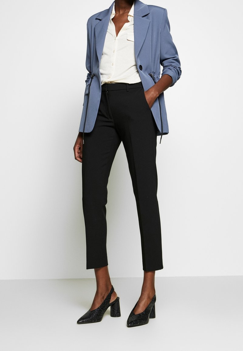 Lichtblauwe getailleerde blazer, wit overhemd met knopen, zwarte op maat gemaakte broek en zwarte schoenen met hak en een textuurlijke afwerking. Strakke lijnen en moderne snit.