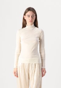 Maglione beige chiaro a maniche lunghe con un colletto a polo arricciato, realizzato in un materiale trasparente e morbido, abbinato a pantaloni a gamba larga lucidi e paillettati.
