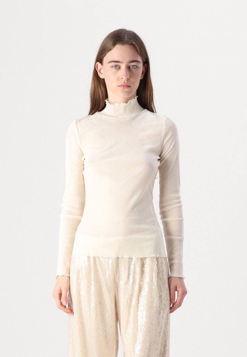 Maglione beige chiaro a maniche lunghe con un colletto a polo arricciato, realizzato in un materiale trasparente e morbido, abbinato a pantaloni a gamba larga lucidi e paillettati.