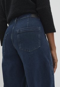 Jeans in denim blu scuro con vita alta, dotati di due tasche posteriori e dettagli di cucitura minimali. Il tessuto ha una texture liscia.