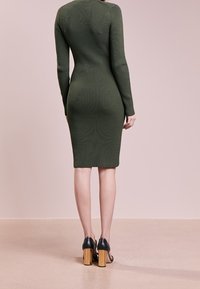 Donna che indossa un abito midi verde oliva a maniche lunghe e righe, con tacchi aperti neri e tacco in legno, in piedi con il viso rivolto verso il lato opposto.