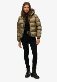 Olijfgroen pufferjack met een capuchon, ritsen en elastische manchetten, gedragen over een zwarte gebreide top en gecombineerd met zwarte skinny jeans en veterschoenen.