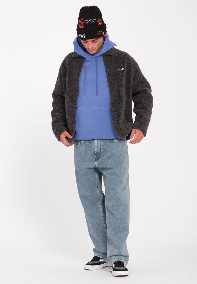 Giacca in pile grigia sopra un felpa blu, jeans azzurri chiari e sneakers nere. Berretto nero con grafiche colorate e giocose.