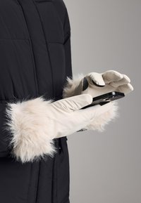 Beige handskar med mjuka fuskpälsmanschetter, båda används medan de håller en svart smartphone. Handskarna har en jämn yta och en figurnära design.