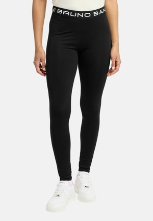 Person trägt schwarze Slim-Fit-Leggings mit gebrandetem Taillenbund und weißen Sneakers, steht vor einem einfarbigen hellen Hintergrund.
