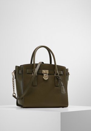Sac à main en cuir vert olive avec doubles poignées, une bandoulière amovible et un charm fermeture doré sur un piédestal blanc.