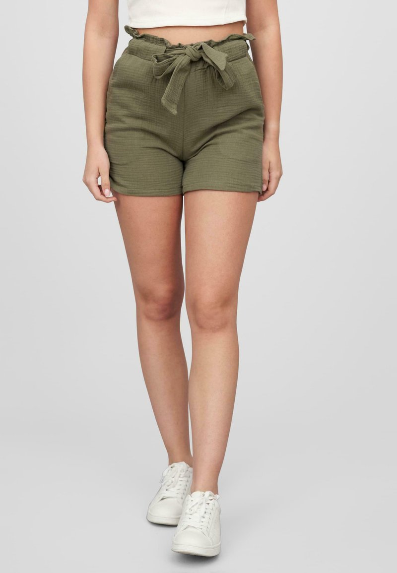 Olivengrønne shorts med struktureret stof, høj talje, flæseliv og bælte med snøre, parret med hvide sneakers.