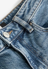 Ljust blå denimjeans med en synlig dragkedja och metallknapp, med kontrasterande sömmar och en texturerad tyg. Detalj av midjebandet inkluderat.