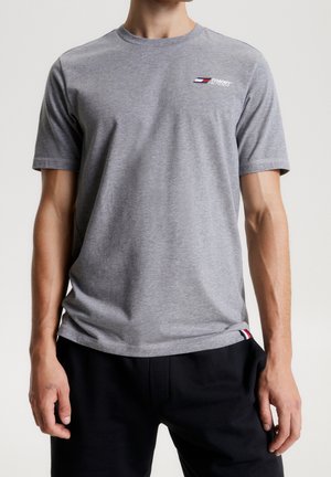 Homme portant un t-shirt gris ajusté à manches courtes Tommy Hilfiger et un pantalon noir, debout les bras détendus le long du corps.