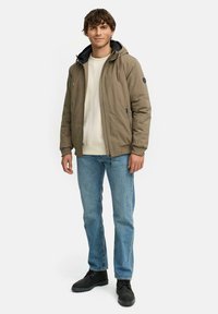 Giacca khaki con cappuccio, polsini elastici, chiusura con zip e due tasche laterali. Indossata sopra un maglione beige chiaro e jeans di denim. Scarpe nere.