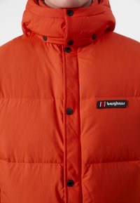 Rød boblejakke med høy krage, med svarte trykknapper og et logo-patch på venstre brystområde. Lett, quiltet tekstur.