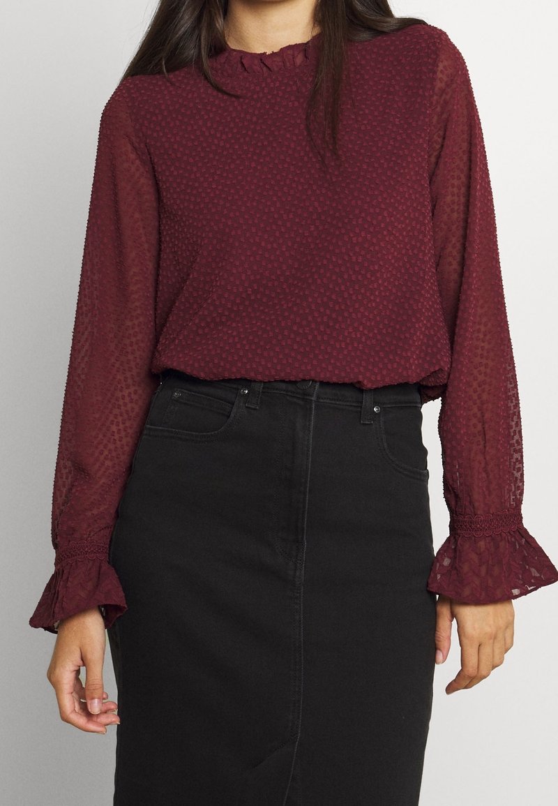 Bordeauxrote transparente Bluse mit strukturiertem Muster, gerafftem Ausschnitt und ausgestellten Manschetten, kombiniert mit einem schwarzen, hoch taillierten Rock.