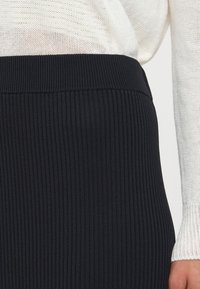 Fustă neagră cu nervuri, având un talie lată și un material texturat. Marginea de jos este netedă, completând un top tricotat de o culoare deschisă.