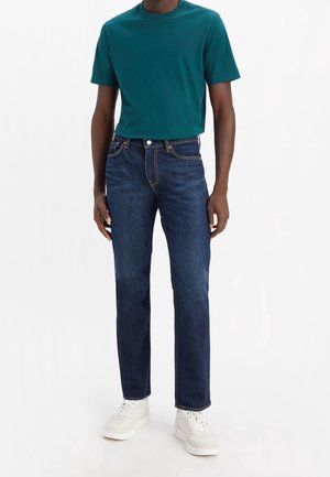Homme portant un jean bleu foncé, un t-shirt turquoise à manches courtes et des baskets blanches, debout devant un fond blanc.