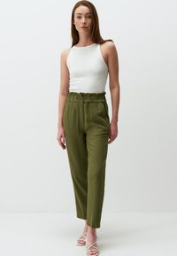 Witte geribde tanktop gecombineerd met hoogzitende olijfgroene broeken met een gerimpelde taille en rechte pijpen. Weergegeven tegen een effen achtergrond.