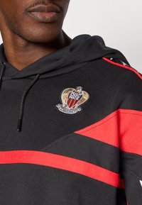 Črna kapa s kapuco z rdečimi črtami, s šivanim logotipom OGC Nice z zlatimi poudarki. Teksturirana tkanina in detajl kapuce z vrvico za zategovanje.