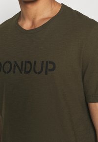 Camiseta de algodón verde oliva con cuello redondo, que presenta un logo negro impreso "DONDUP" en el pecho. Textura suave, manga corta.