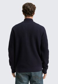 Mornarska pletena pulover s visokim ovratnikom in dolgimi rokavi, prikazan zadaj. Uporabljen v kombinaciji s svetlo modrimi jeans hlačami z detajli šivanja.