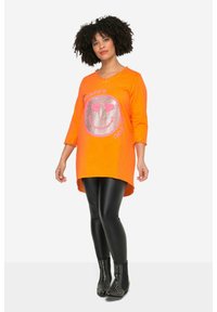 Orangene Tunika mit silbernem, glitzerndem Smiley-Gesicht, schwarze Lederleggings und Nieten-Stiefeletten. Hat eine lockere Passform und 3/4-Ärmel.