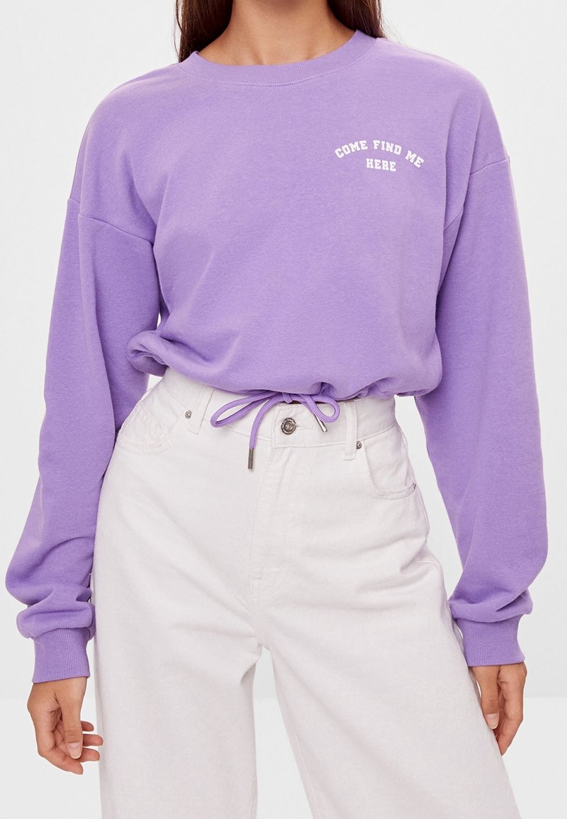 Femme portant un haut à manches longues violet avec le texte « COME FIND ME HERE » et un pantalon blanc taille haute avec un cordon à la taille.